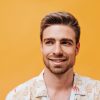 Portrait of nice young man with stylish hairstyle, blue eyes and beard in light cool shirt looking away and smiling on orange backdrop..